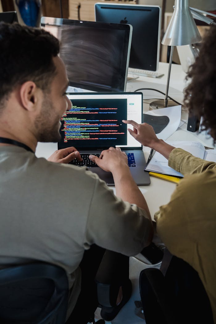 Two programmers working together on a laptop, discussing code in a modern office setting.